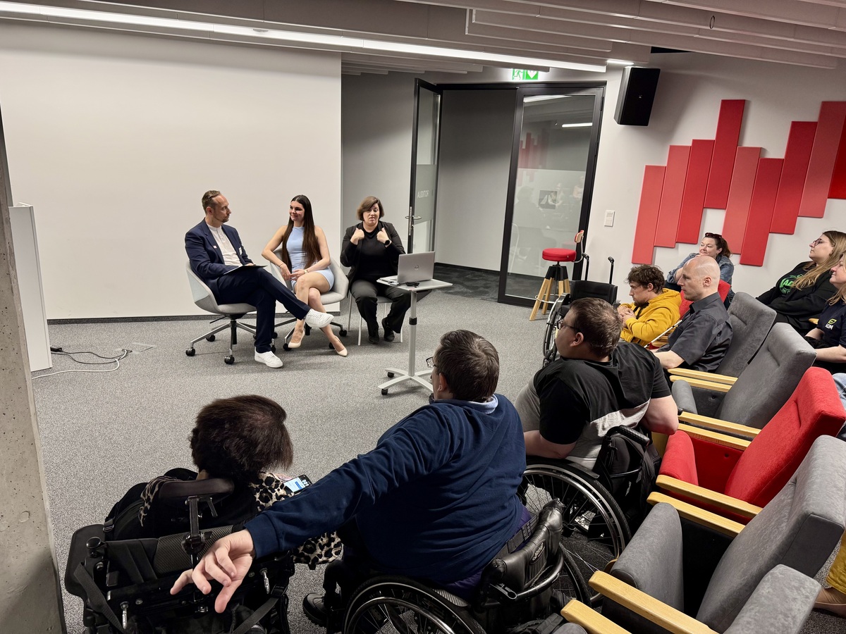 An interview with Krystyna Włodarek and Michał Lisiak in the auditorium. In the foreground, people using wheelchairs. A Polish Sign Language interpreter is in the centre. Open space and participants visible in the background.